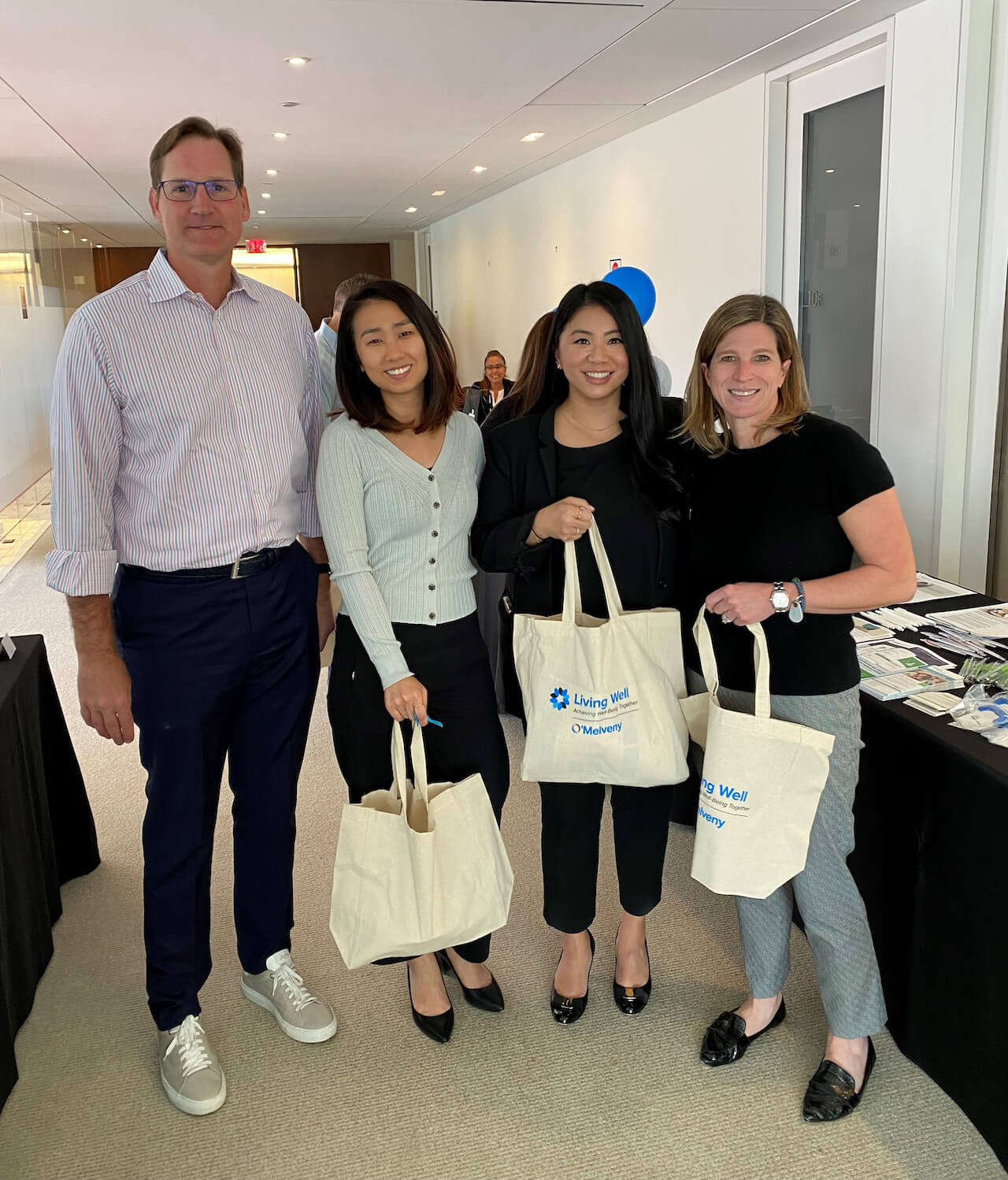 Four colleagues attending a Wellbeing Fair in their office, holding O'Melveny Living Well tote bags.