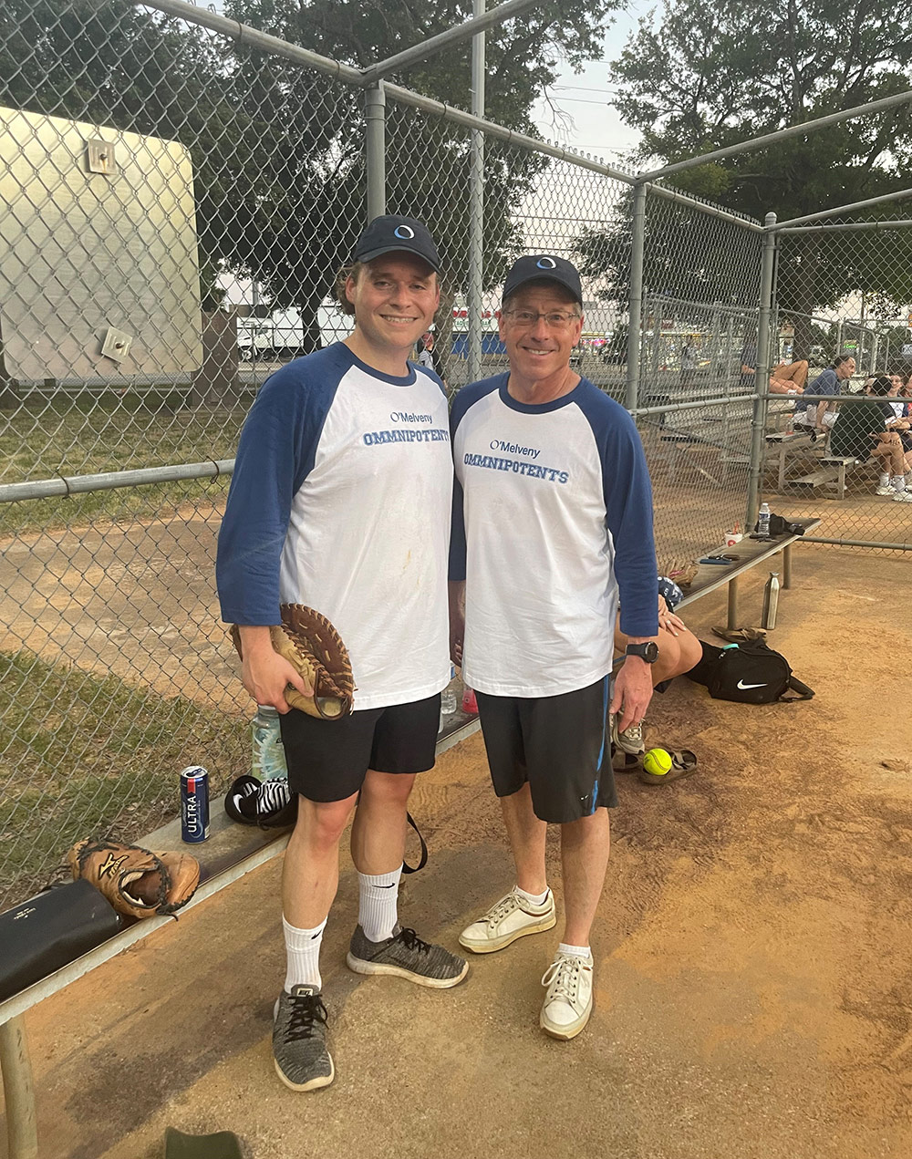 Two colleagues in blue and white O'Melveny t-shirts and hats at a firm baseball game. One colleague is holding a catcher's mitt.